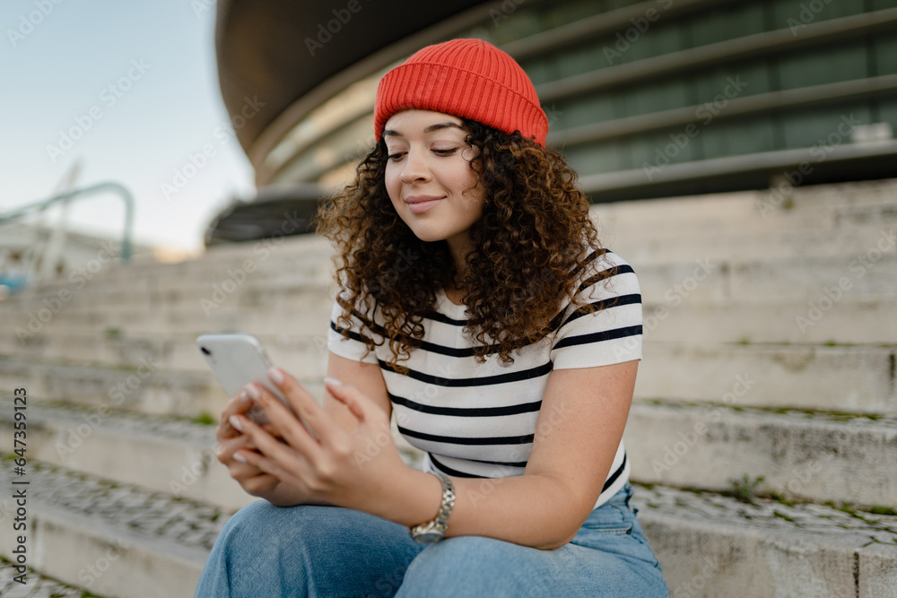 pretty curly smiling woman sitting in city street in striped t-shirt and knitted red hat, using smartphone