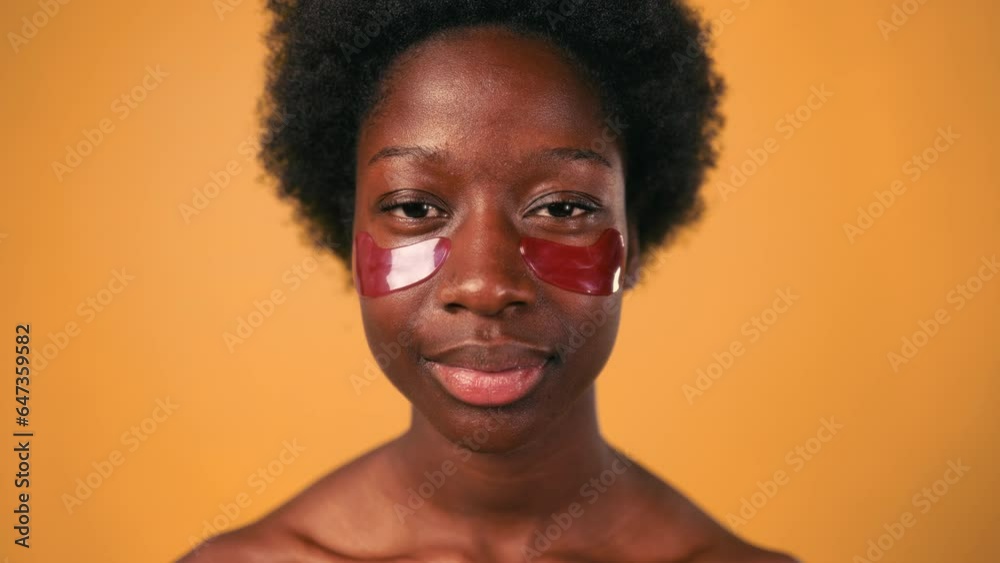 African American young woman with afro hair doing her morning routine using moisturizing eye patches isolated on orange background. Skin care and spa treatments.