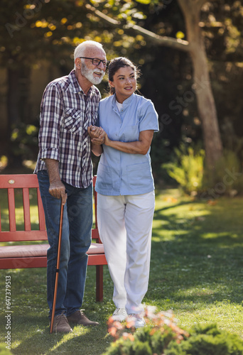 Caregiver helping senior man to walk with stick outdoors