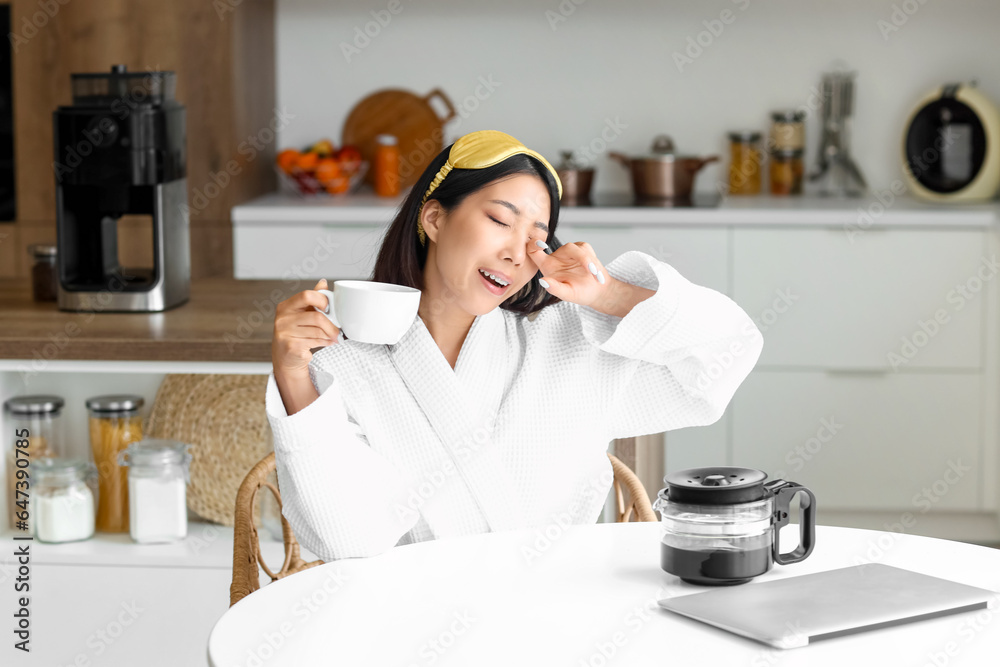 Sleepy Asian woman with cup of hot coffee in kitchen