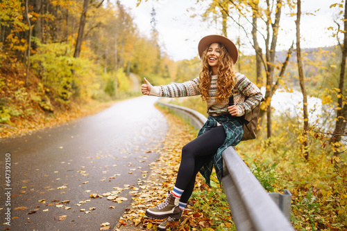 Photography Woman traveler in hiking clothes with a backpack and a hat stands on the side of the road, trying to catch a car on the road