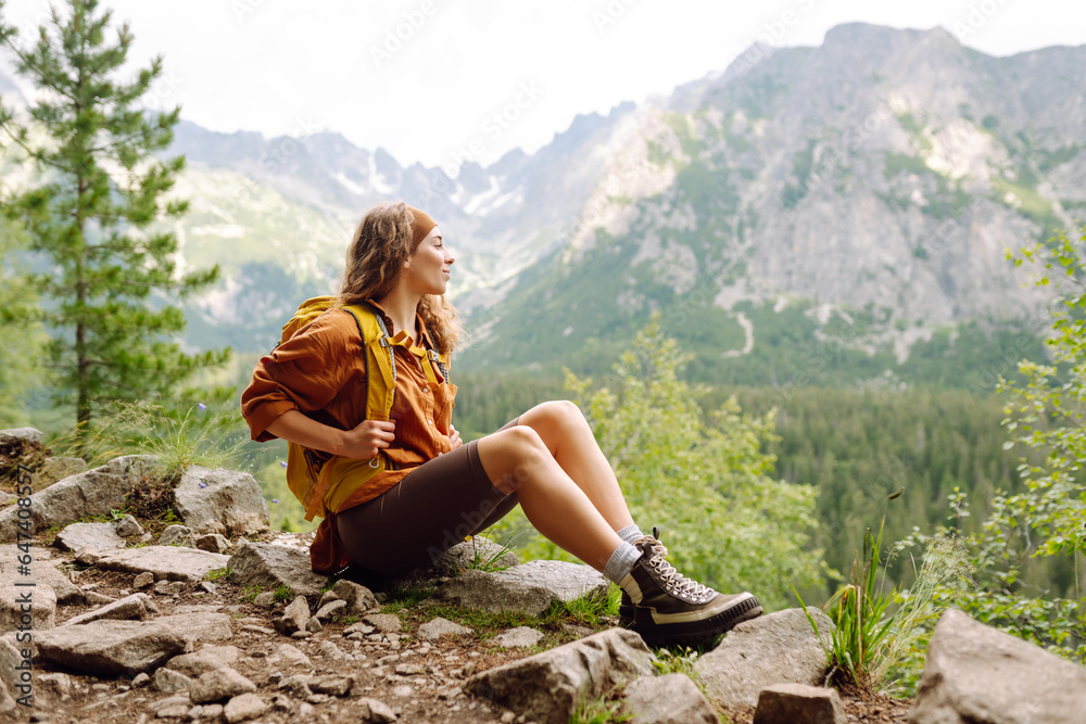 Naklejka premium Active woman with a yellow hiking backpack traveling along hiking trails in the mountains among forests and cliffs. Traveler enjoying nature. Concept of trekking, active lifestyle. Adventures.