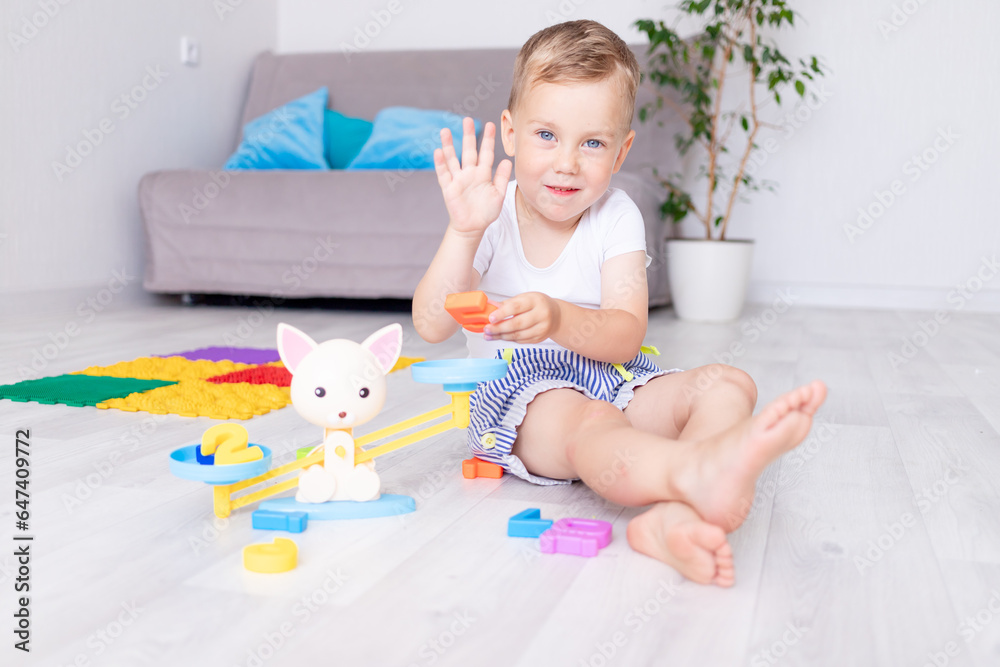 cute baby boy plays with scales at home on the floor and learns to count by showing five fingers on his hand, the concept of early development of children cute baby boy plays with scales at home on the floor and learns to count by showing five fingers on his hand, the concept of early development of children