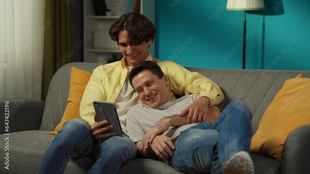 Shot of a homosexual couple at home. They are laying on the couch, watching photos or videos on a tablet, hugging, laughing and expressing love to each other.