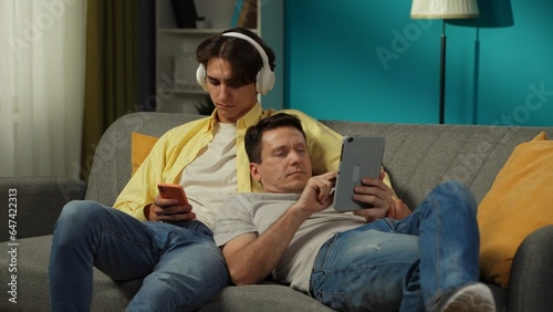 Shot of a homosexual couple at home. They are laying on the couch, one of them is listening to music, the other is scrolling, surfing online on tablet.