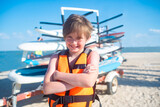 smiling child in a t-shirt with a backpack at the surf station on the beach in summer. joyful 10 year old child stands next to the windsurf boards