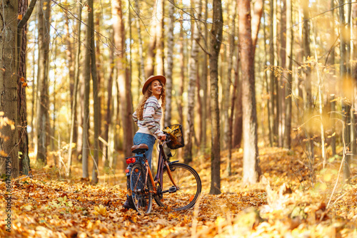 A beautiful woman in stylish clothes and a hat walks and rides a bike in a sunny autumn park. Concept of nature, relaxation. Lifestyle.