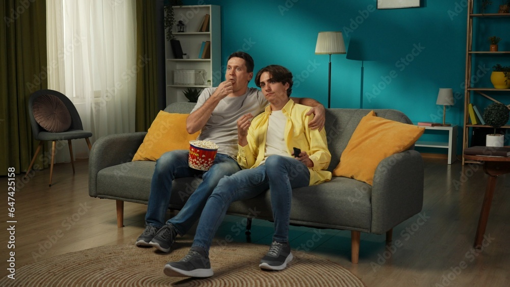 Naklejka premium Full-size shot of a homosexual couple at home. They are sitting on the couch, watching TV together, eating popcorn, hugging, smiling and expressing love to each other.