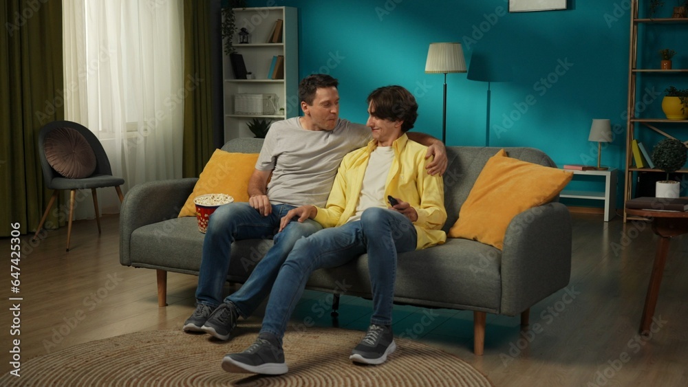 Full-size shot of a homosexual couple at home. They are sitting on the couch, watching TV together, eating popcorn, hugging, smiling and expressing love to each other.
