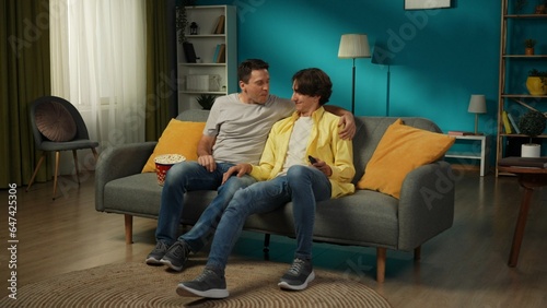 Full-size shot of a homosexual couple at home. They are sitting on the couch, watching TV together, eating popcorn, hugging, smiling and expressing love to each other.