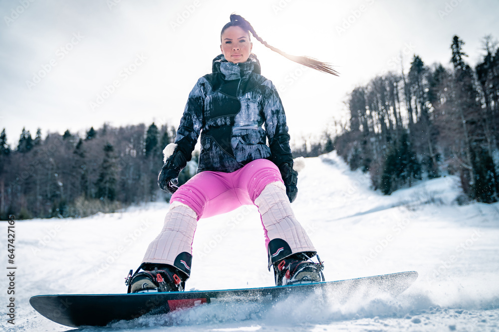 Young female snowboarder having fun on ski track. Fashionable athletic woman wearing pink ski suit and braid showing her sport skills.