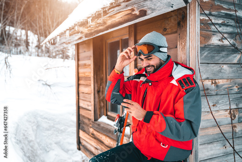 Young smiling male skier using smart phone in front of log cabin. Fashionable man in full ski equipment checking his mobile phone on vacation.