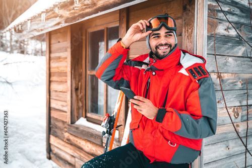 Young smiling male skier using phone in front of log cabin. Happy guy in skiwear taking a break outdoors. Confident man with mobile phone taking ski goggles off and looking away. photo