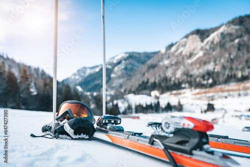Close up of skis, ski goggles, ski gloves and ski poles. Sports equipment displayed in snow with magnificent mountains and clear sky in the background.