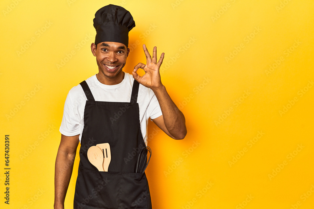 African American cook with utensils, yellow studio, cheerful and confident showing ok gesture.