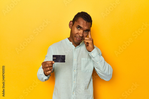African American man with credit card, yellow studio, pointing temple with finger, thinking, focused on a task.