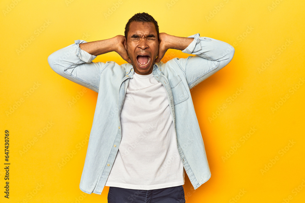 African American man in blue shirt, yellow studio, covering ears with hands trying not to hear too loud sound.