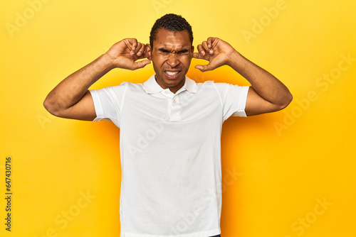 African American man in white polo, yellow studio, covering ears with fingers, stressed and desperate by a loudly ambient.