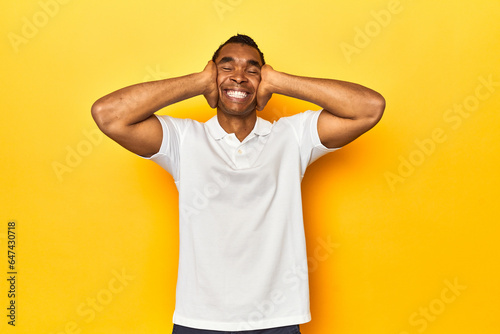 African American man in white polo, yellow studio, laughs joyfully keeping hands on head. Happiness concept.