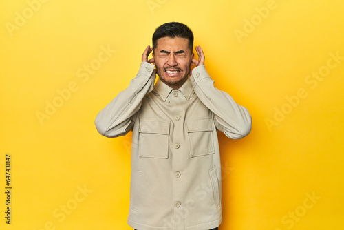 Asian man in stylish gray shirt on yellow studio covering ears with hands.