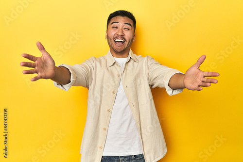 Casual Asian man with open shirt, white tee on yellow studio feels confident giving a hug to the camera.