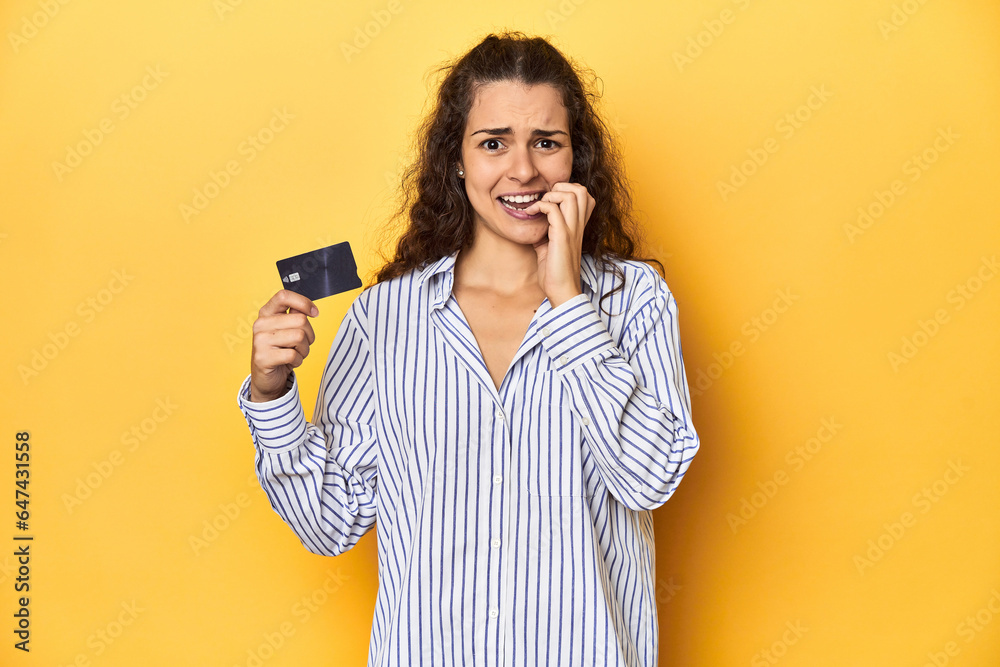 Woman holding a credit card, yellow backdrop, biting fingernails, nervous and very anxious. Woman holding a credit card, yellow backdrop, biting fingernails, nervous and very anxious.