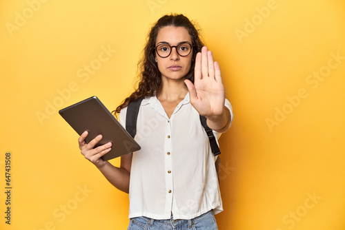 Student holding a tablet, glasses, backpack on, standing with outstretched hand showing stop sign, preventing you.