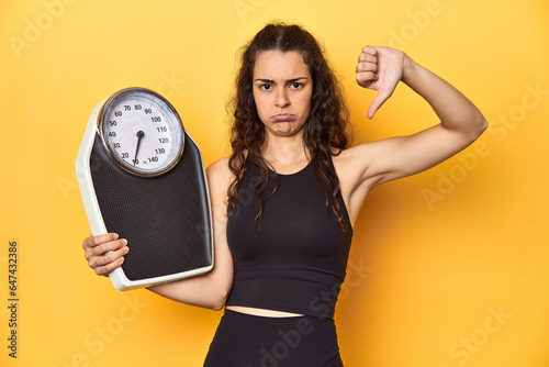 Worried young Caucasian woman in sportswear holding a scale, fitness concept.