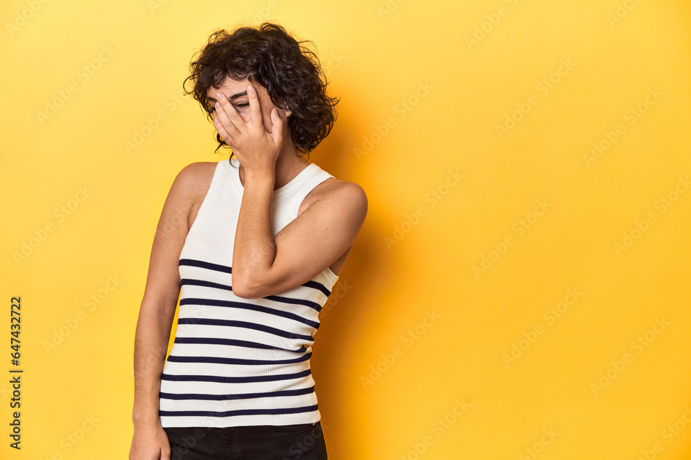 Caucasian curly-haired woman in white tank-top blink at the camera through fingers, embarrassed covering face. Caucasian curly-haired woman in white tank-top blink at the camera through fingers, embarrassed covering face.