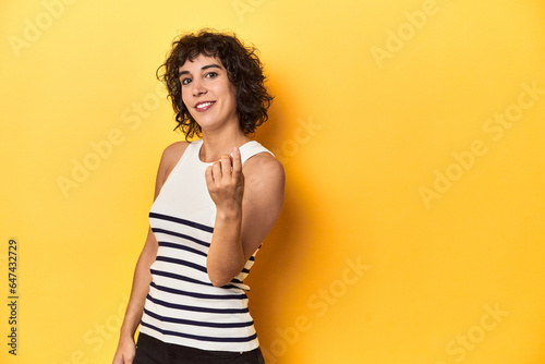 Caucasian curly-haired woman in white tank-top pointing with finger at you as if inviting come closer.