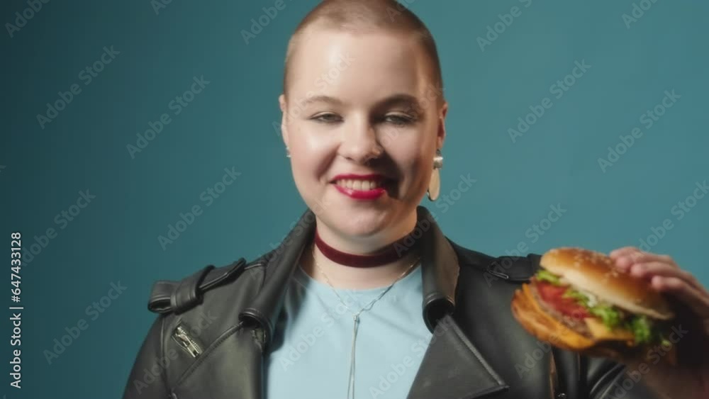 Chest up portrait of young Caucasian body positive woman with shaved head and red lips posing for camera with delicious burger in hand standing on turquoise background Chest up portrait of young Caucasian body positive woman with shaved head and red lips posing for camera with delicious burger in hand standing on turquoise background