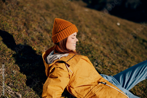 Woman beautifully lying on a hill on the grass holiday smile with teeth in the mountains in the autumn in a yellow raincoat and jeans happy sunset trip on a hike, freedom lifestyle 