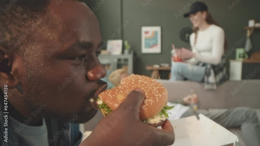 Side view medium closeup of African American guy taking bite of delicious saucy burger while chilling with company of diverse friends at home on weekend