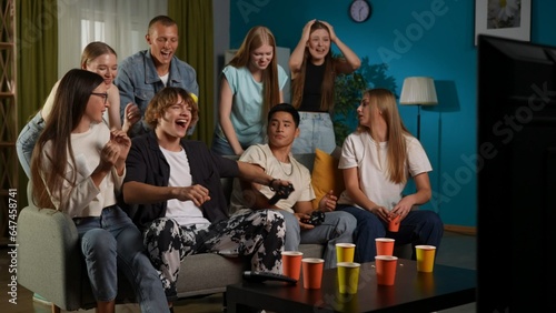 Group of teens, young people, friends sitting and standing around two guys, boys who were playing a console, gaming. One of them is celebrating his win emotionally. Full-size.