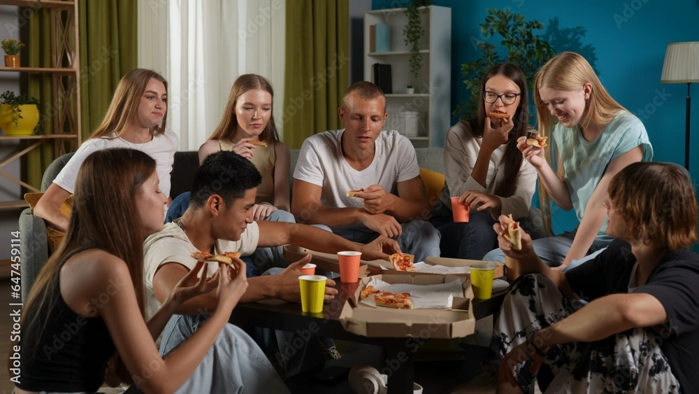 Medium shot of a group of teens, young people, friends sitting on a couch and floor around the table, eating pizza, drinking beverages, chatting, talking, joking around.