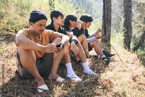 Young hiker man using binocular with group of multiethnic friends hanging out together sitting in a row at the top of the hill