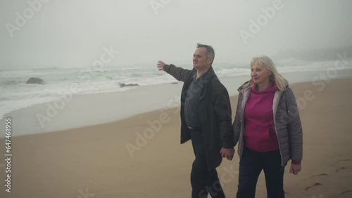 Tracking shot of a senior Caucasian White couple walking on the beach holding hands and chatting with stormy ocean in the background photo