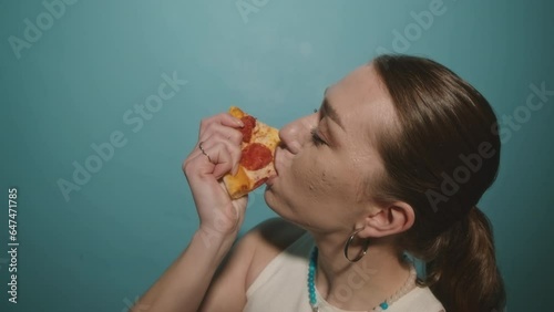 Chest up of hungry Caucasian girl in her early twenties eating delicious pepperoni with pleasure standing on turquoise background