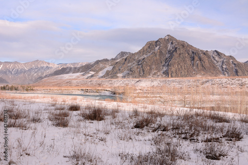 A flat snow-covered valley at the foot of a high mountain is crossed by a beautiful winding river on a sunny winter day.