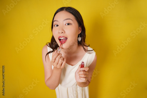 Asian young woman applies lipstick on lips. Portrait of beautiful glamor model with stylish makeup on the face. Asian girl with fashion make up on yellow background. Doing cosmetic makeup tutorial.
