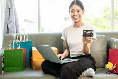 Asian woman holding credit card and smart phone and laptop sitting on sofa at home doing online banking transactions in online shopping
