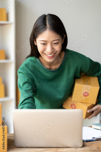 Portrait of a young Asian woman Ecommerce sitting in a home office full of parcels in the background Write order notes with a laptop and calculator for e-commerce and SME shipping businesses. © ArLawKa