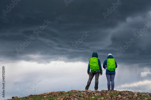 Close-up view, taken from behind, of two women wearing backpacks standing on a mountain ridge under a dark and foreboding sky near Whitehorse; Whitehorse, Yukon Territory, Canada photo
