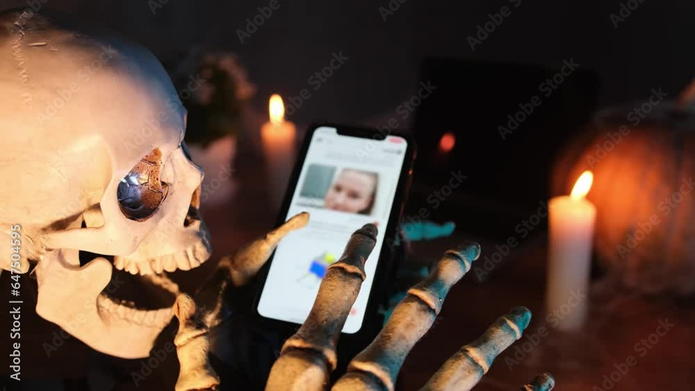 Side view, passionately skeleton scrolls through feed on smartphone screen with bone fingers in darkness against the backdrop of burning candle and pumpkin, Happy Halloween attributes. Close up.