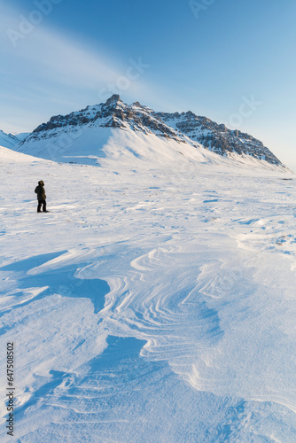 Man In A Parka Standing On The Windswept Snow, Sastrugi Patterns In The Foreground, Soakpak Mountain In The Distance, Anaktuvuk Pass, Gates Of The Arctic National Park; Alaska, United States Of Americ photo