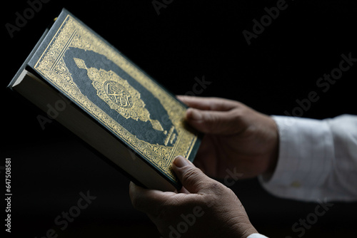 A Muslim man holds a Quran in his hand as the mosque lights shine through the window.
