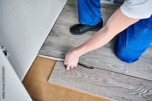Close up of male construction worker hand using tape measure while measuring distance from wall to laminate board. Man installing laminate flooring in apartment under renovation.