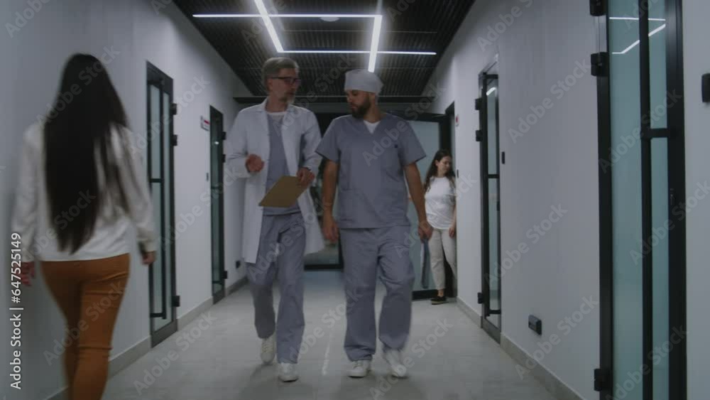 Mature doctor with colleague walk together the clinic corridor. Healthcare professionals discuss treatment, diagnosis or medical test results. Medical staff and patients in hospital or medical center. Mature doctor with colleague walk together the clinic corridor. Healthcare professionals discuss treatment, diagnosis or medical test results. Medical staff and patients in hospital or medical center.