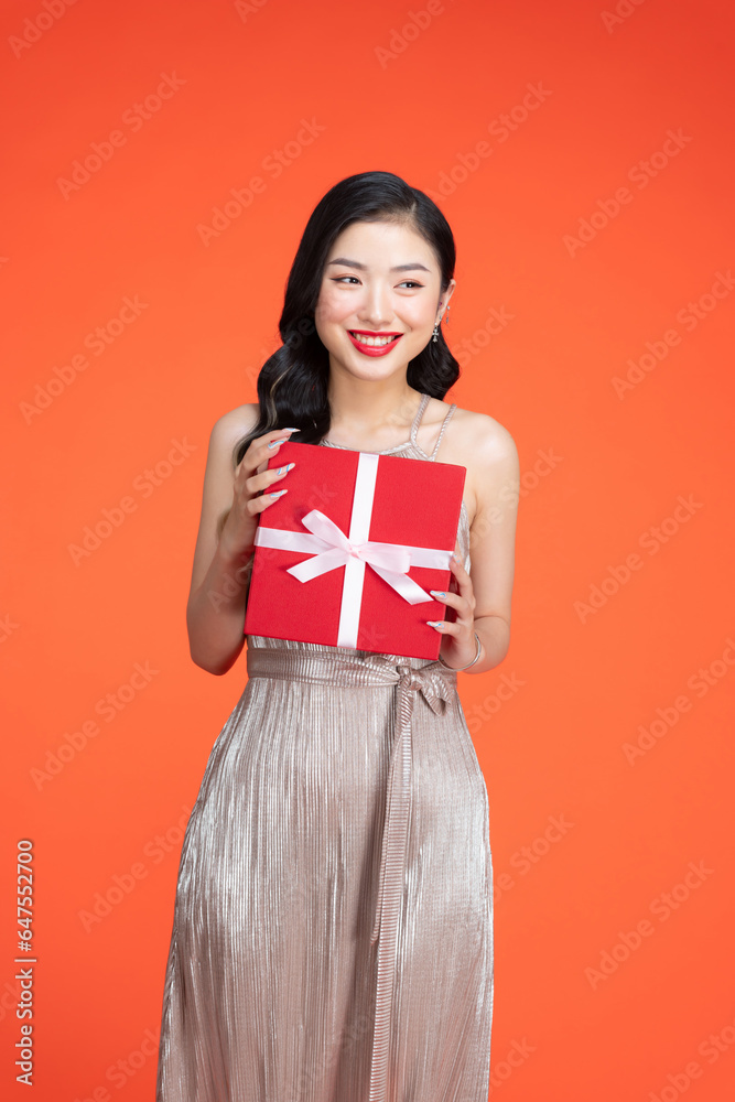 Fototapeta premium Young woman wearing a shiny dress holding a red gift box in front of a red backdrop