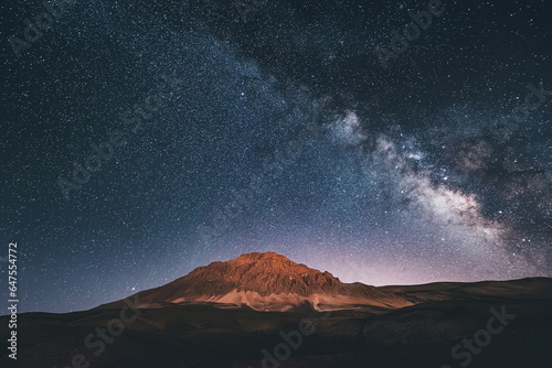 Amidst the serene mountains, the milky way paints a cosmic masterpiece against the deep blue night sky, creating a breathtaking scene. © EdNurg